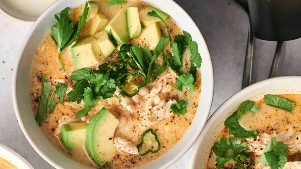 Close-up of a bowl of creamy soup garnished with avocado slices, cilantro, and sliced chili peppers.