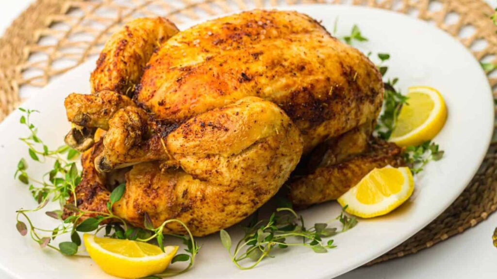 A roasted chicken garnished with lemon slices and herbs on a white plate.
