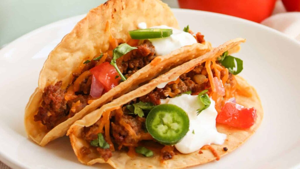 Two crispy tacos filled with ground beef, diced tomatoes, shredded cheese, sliced jalapeños, sour cream, and cilantro on a white plate.