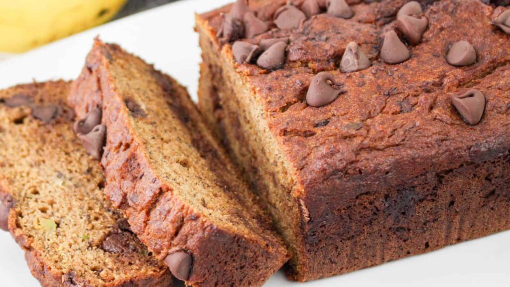 A loaf of chocolate chip banana bread is sliced, showing a dense texture with visible banana and chocolate chip pieces on the surface and inside.