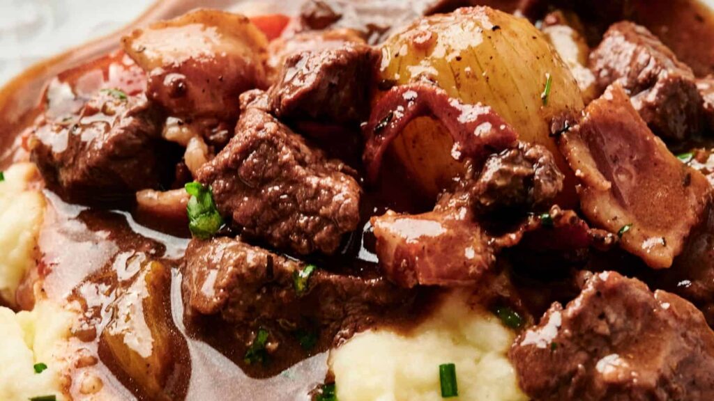 Close-up of a hearty beef stew with pieces of beef, bacon, onions, and a rich gravy, served over a bed of creamy mashed potatoes.