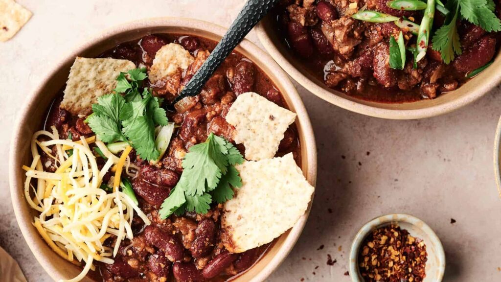 Two bowls of chili with kidney beans, topped with cheese, cilantro, tortilla chips, and sliced green onions. A spoon is placed in one bowl. A small dish of chili flakes is nearby.