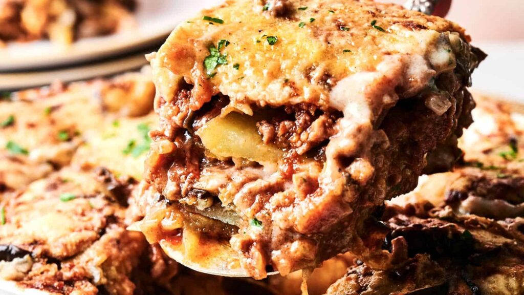 Close-up of a serving of moussaka on a spatula. Layers of eggplant, meat, and cheese are visible, topped with melted cheese and garnished with parsley.