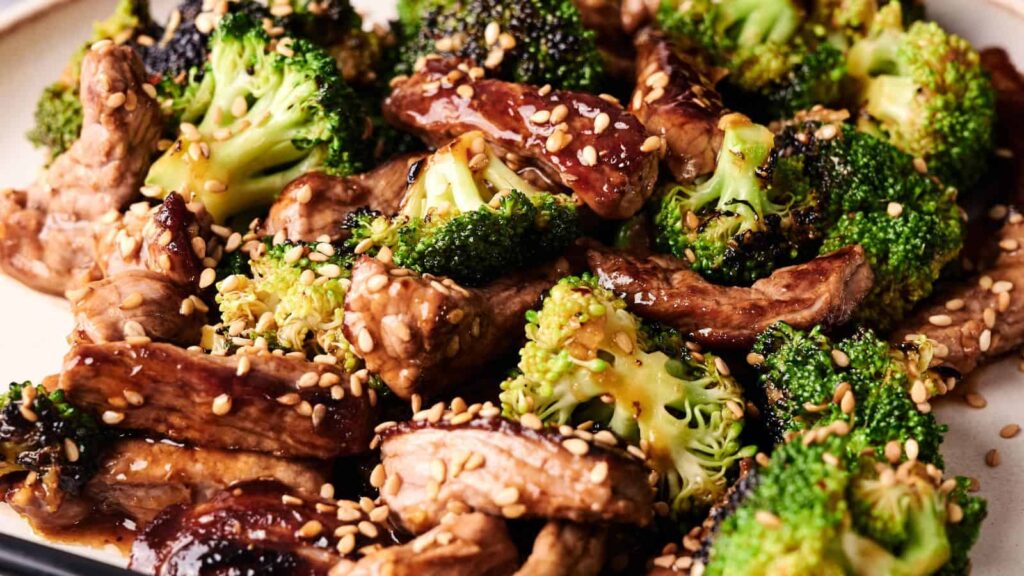Close-up of a beef and broccoli stir-fry topped with sesame seeds, featuring cooked beef strips, broccoli florets, and a glossy sauce.