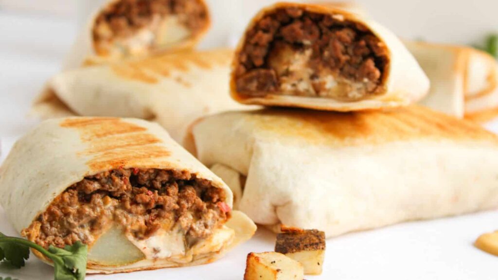 Grilled beef burritos with creamy filling, arranged on a table, some cut open to show cross-section of ingredients. Pieces of diced potato and cilantro garnish are visible nearby.