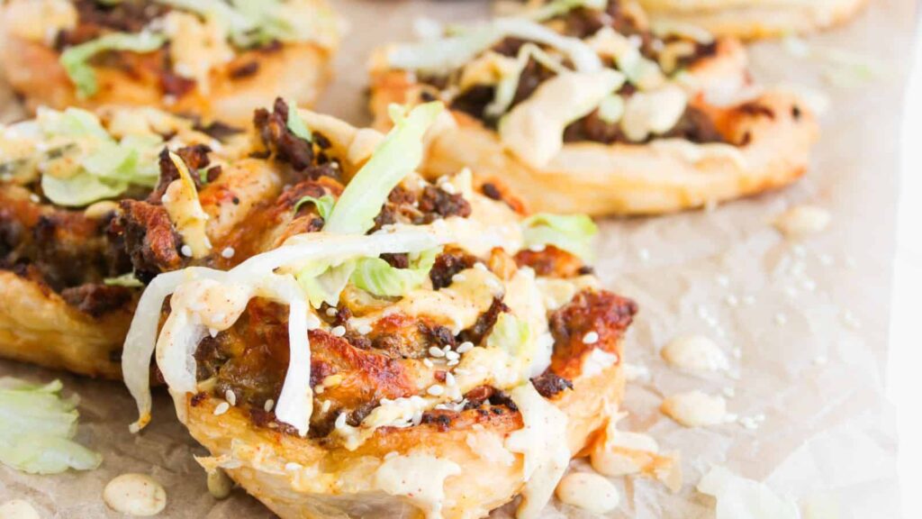 Close-up of puff pastry rounds topped with ground meat, shredded lettuce, sesame seeds, melted cheese, and creamy sauce on parchment paper.
