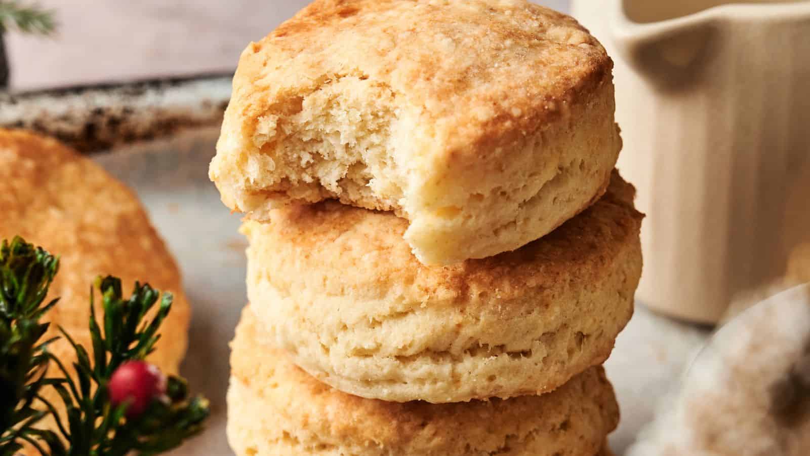 Three golden-brown biscuits are stacked, with the top one missing a bite. A small sprig of greenery with red berries is visible in the corner.