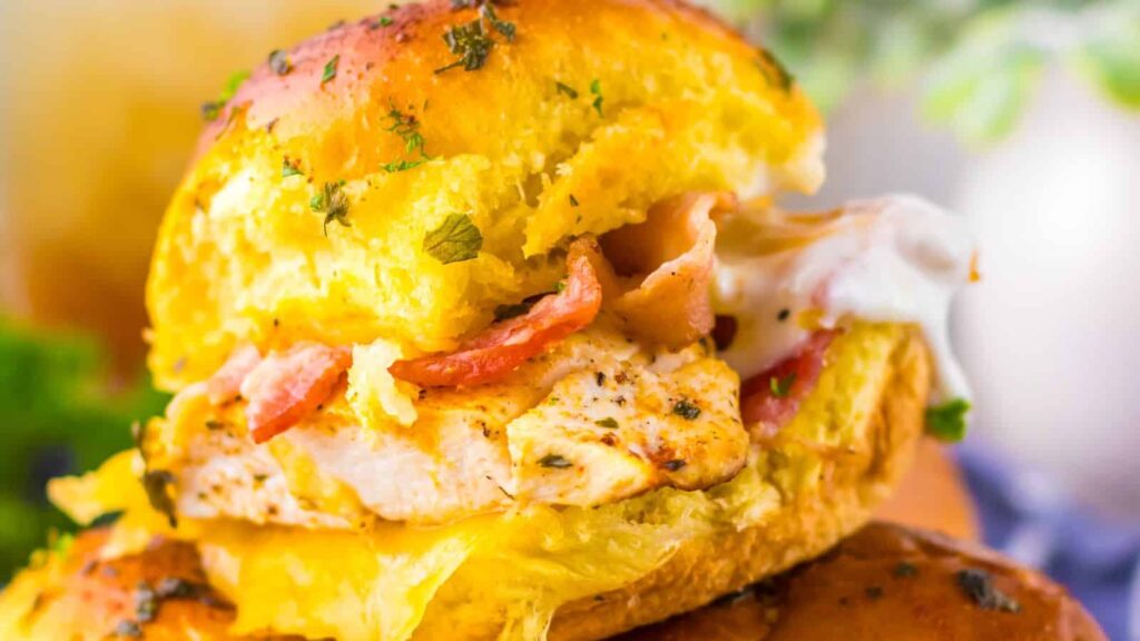 A close-up of a chicken sandwich with bacon, melted cheese, and herbs on a toasted bun.