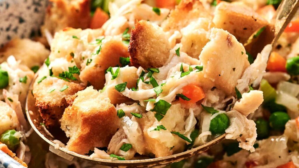 Close-up of a spoon scooping savory stuffing with chunks of bread, peas, carrots, and herbs.