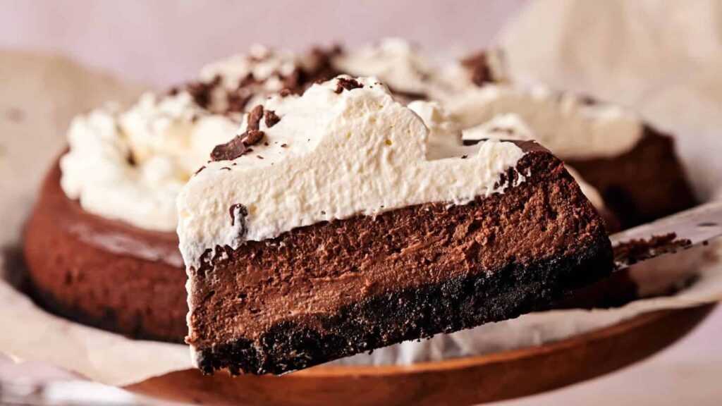 A slice of chocolate cheesecake with a dark crust and whipped cream topping being lifted from the whole cake.
