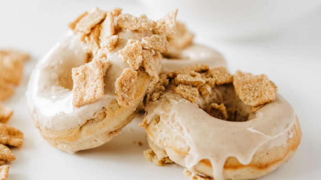 Two glazed donuts topped with crumbled cereal pieces are set on a white surface.