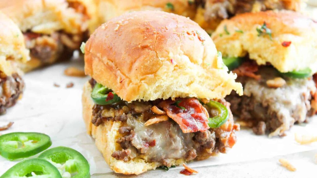 A close-up of a slider sandwich with ground beef, melted cheese, bacon, and jalapeño slices on a soft bun, with more sliders and jalapeño slices in the background.
