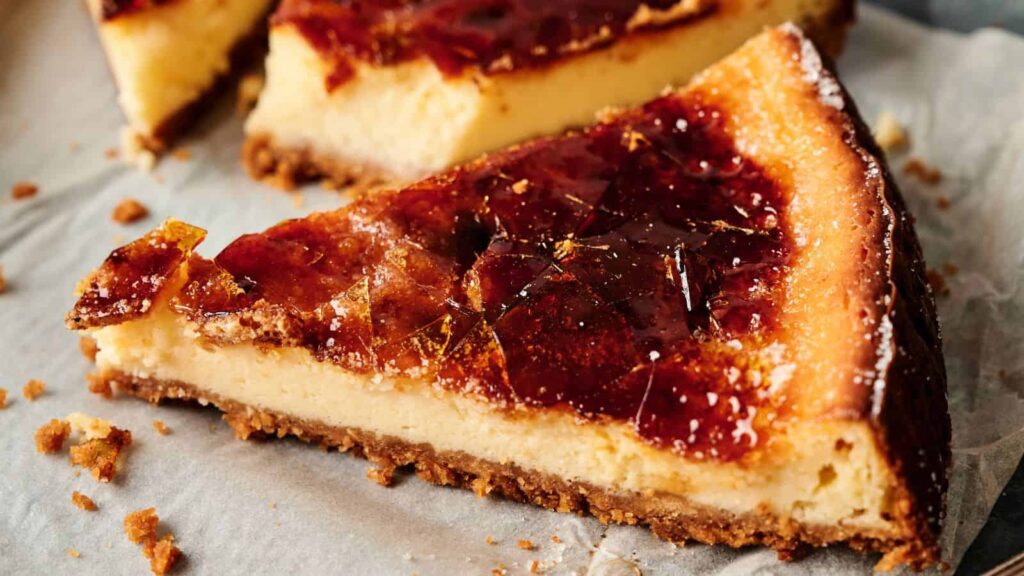 A close-up of a slice of cheesecake with a caramelized sugar topping, resting on parchment paper with a crumbly base visible.
