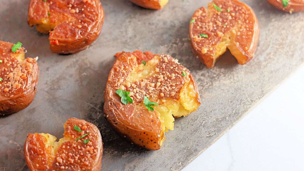 Several smashed roasted potatoes topped with grated cheese and chopped parsley are arranged on a metal baking sheet.