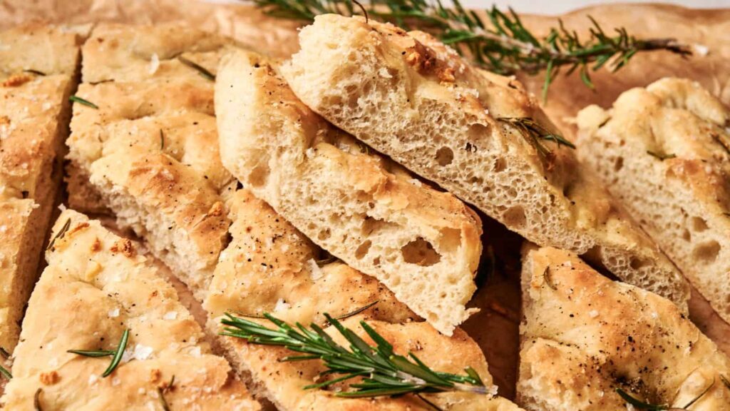 Slices of focaccia bread garnished with rosemary sprigs are stacked on parchment paper.