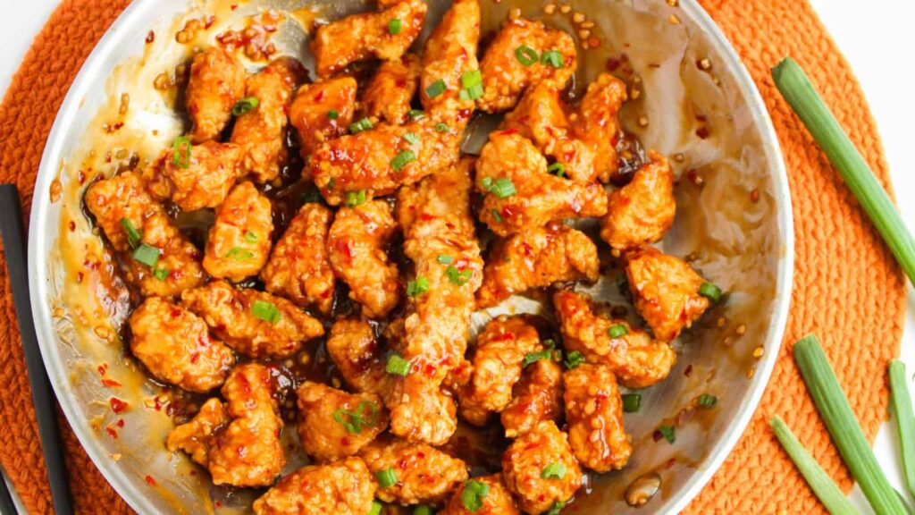 A skillet filled with crispy chicken pieces coated in a sticky, brown sauce, garnished with chopped green onions, placed on an orange woven mat with green onion stalks beside it.
