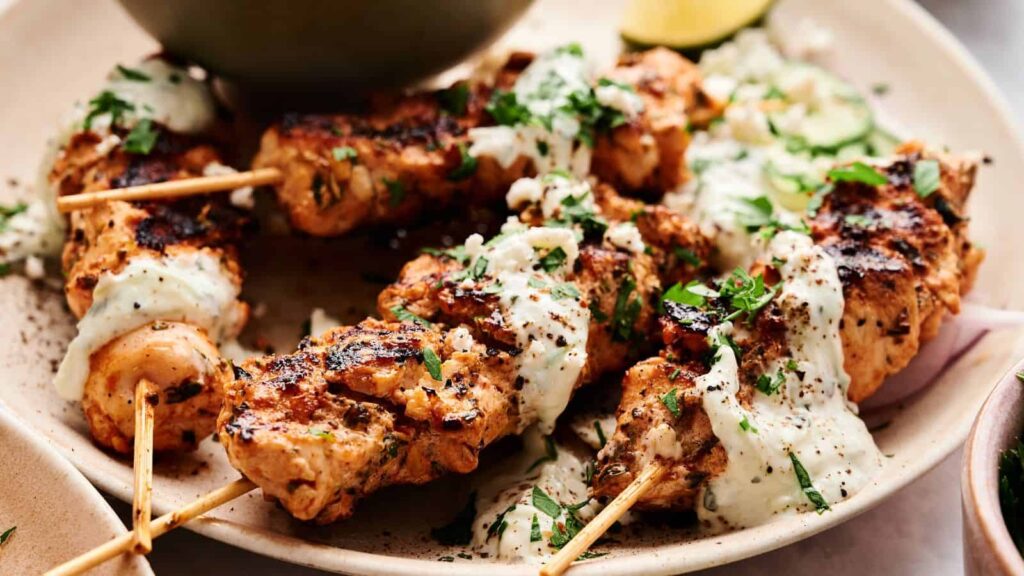 Grilled chicken skewers topped with a creamy white sauce and chopped herbs, served on a plate with sliced cucumbers and onions.