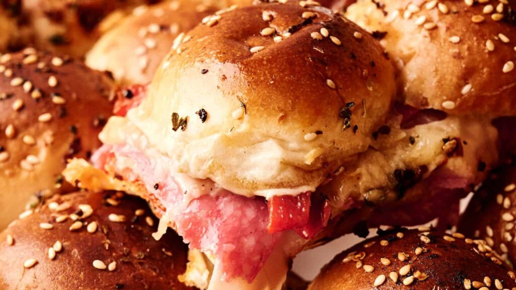 A close-up of a sandwich slider with layers of meat and melted cheese between sesame seed buns.