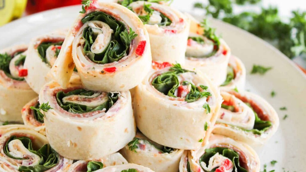 A plate of tortilla pinwheel sandwiches filled with leafy greens, cream cheese, and diced red peppers, garnished with fresh parsley.