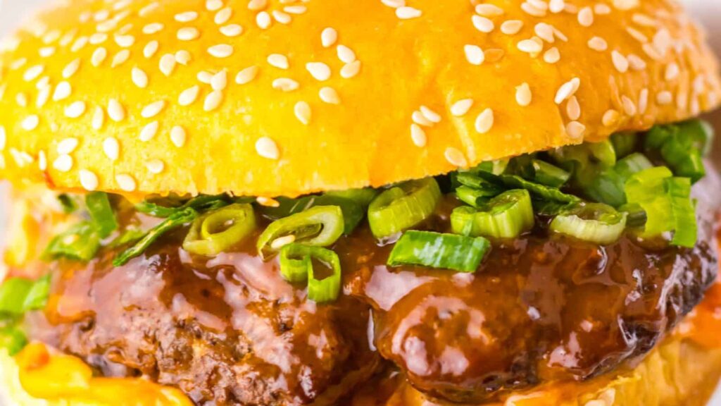 Close-up of a sesame seed bun burger with beef patty, topped with brown sauce and chopped green onions.