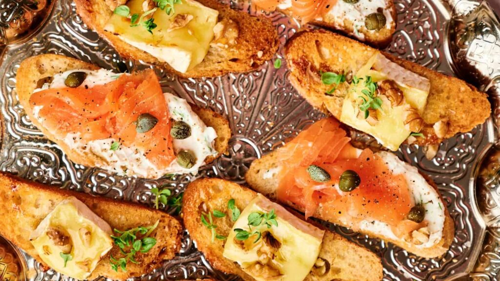 A platter of bruschetta topped with smoked salmon, capers, cheese, walnuts, and herbs on toasted baguette slices.