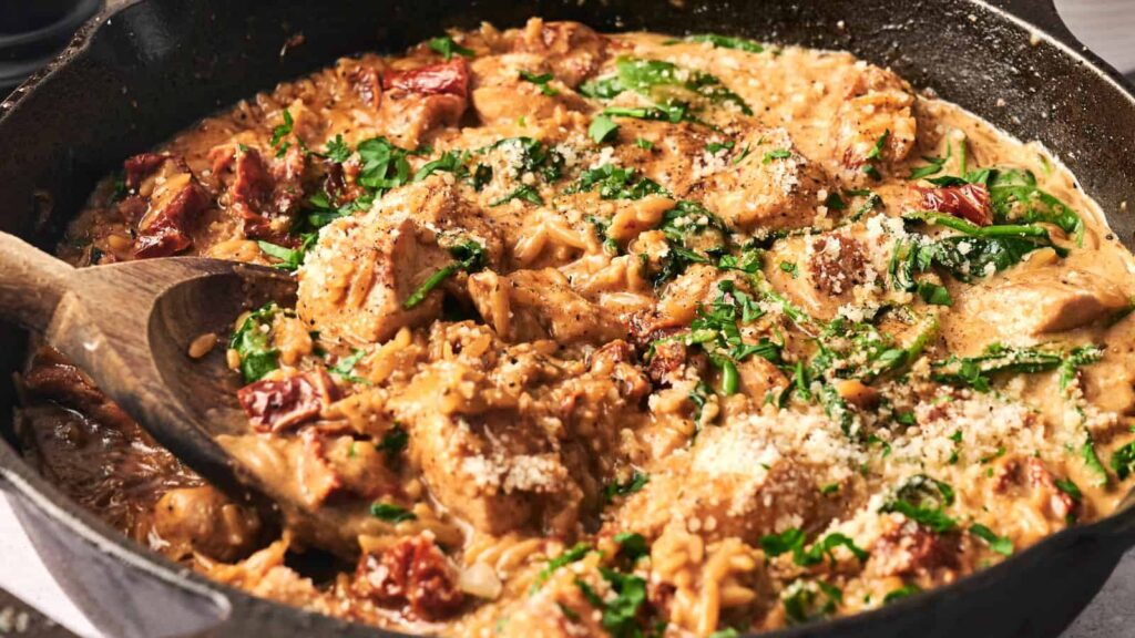 A skillet filled with creamy chicken pasta, featuring chunks of chicken, spinach, sun-dried tomatoes, and a sprinkle of grated cheese, mixed with a wooden spoon.