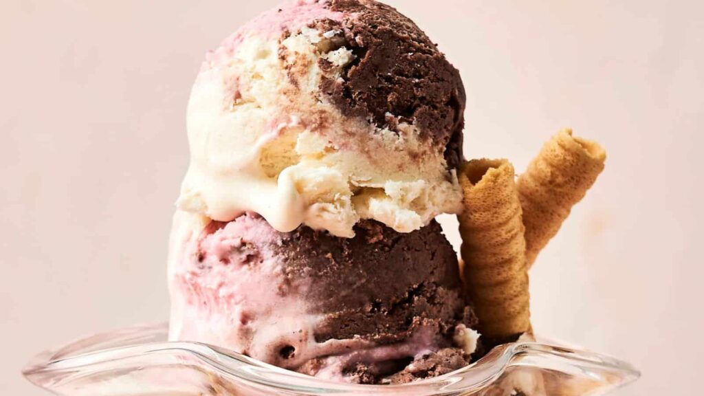 Two scoops of Neapolitan ice cream in a clear glass dish, featuring chocolate, vanilla, and strawberry flavors, with two rolled wafer cookies on the side.