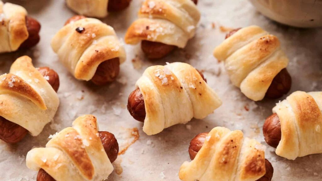 Close-up of multiple pigs in a blanket, which are small sausages wrapped in golden, flaky pastry on a parchment-lined surface, sprinkled with coarse salt.