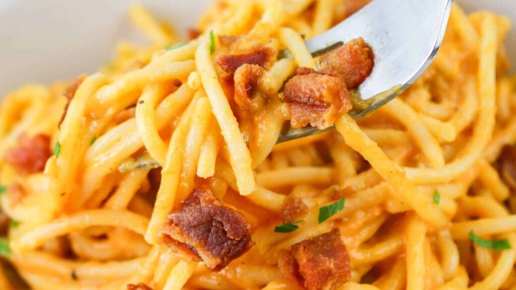 A close-up of a forkful of spaghetti carbonara with crispy bacon bits and garnished with green herbs.