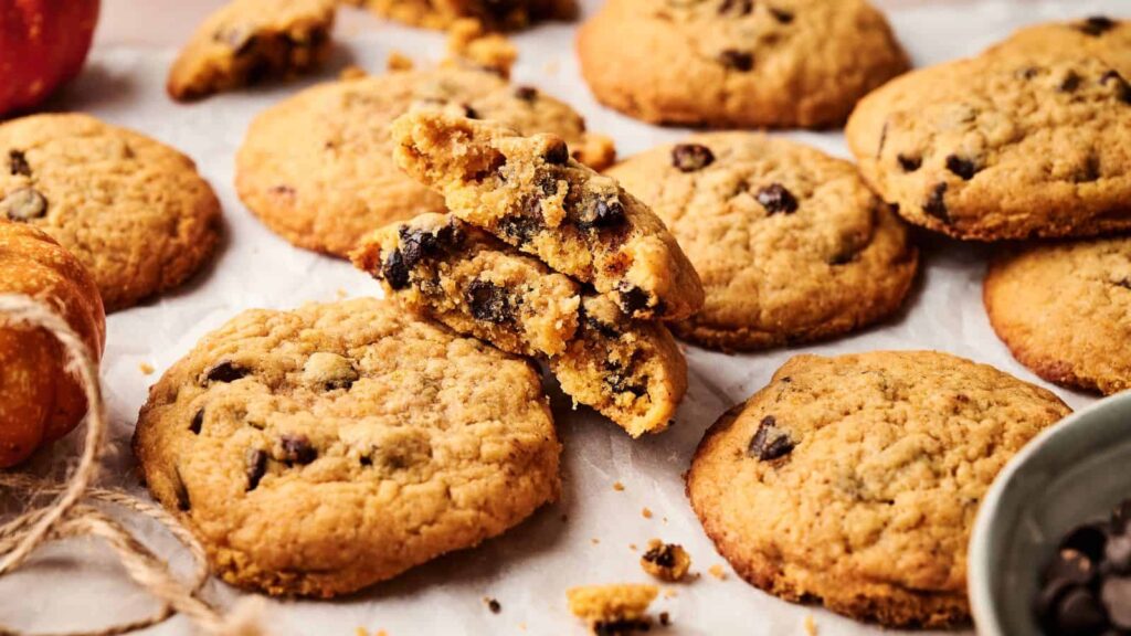 A close-up of several chocolate chip cookies on parchment paper, with one cookie broken in half to show the chocolate chips inside.