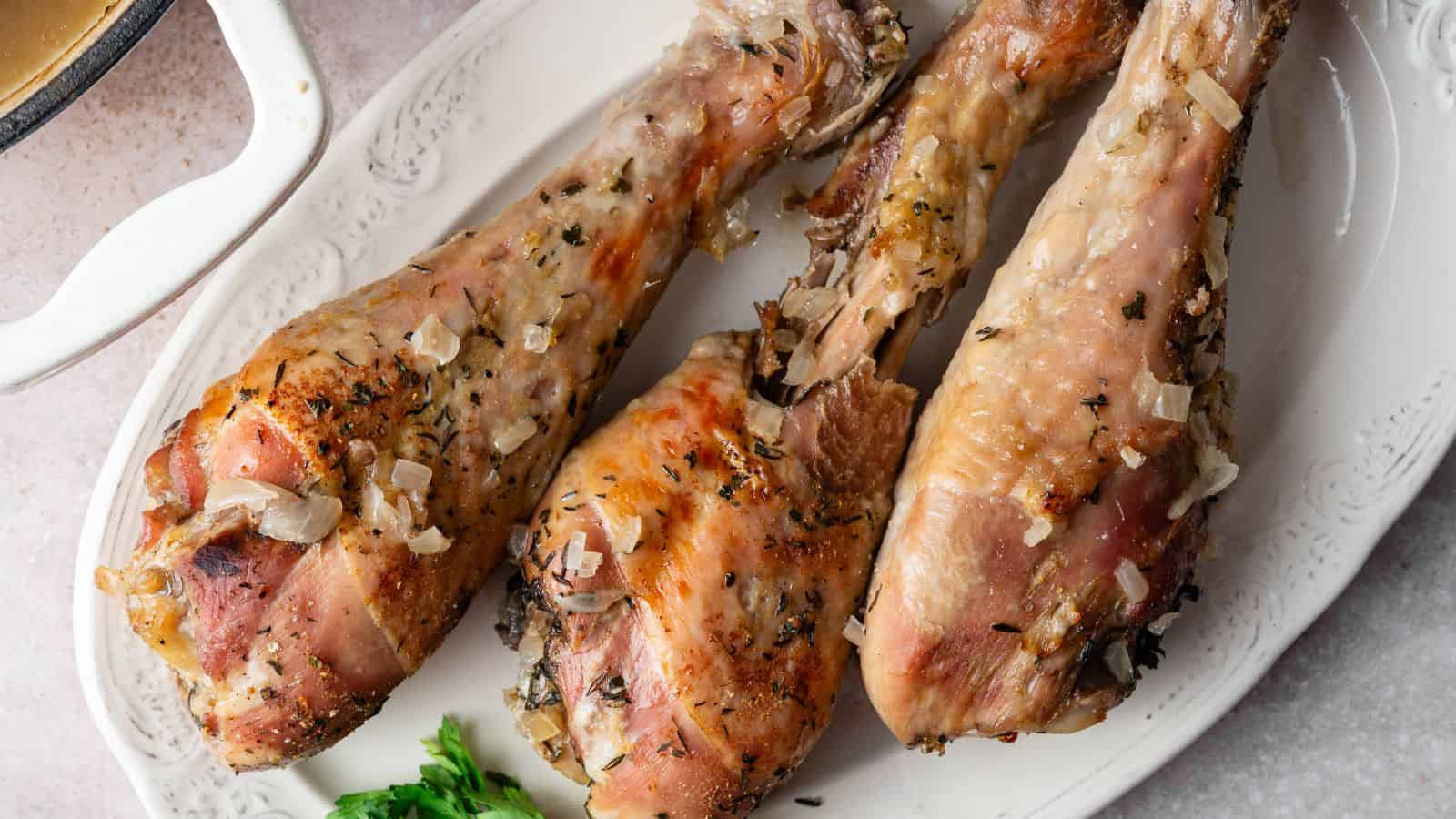 Three cooked turkey legs with herbs and chopped onions are arranged on a white oval platter, garnished with a sprig of parsley.