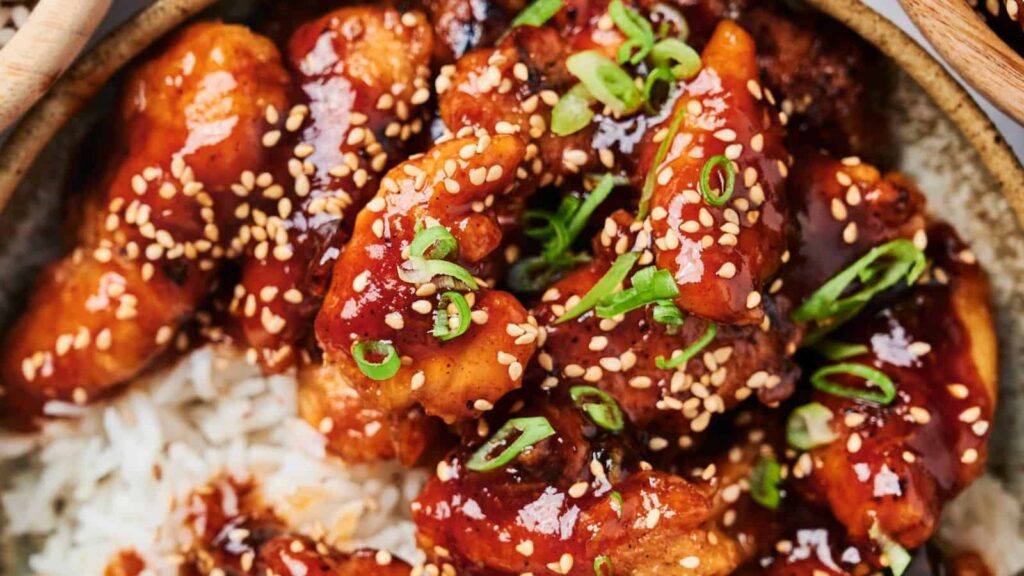Close-up of glazed chicken pieces topped with sesame seeds and chopped green onions, served over a bed of white rice.