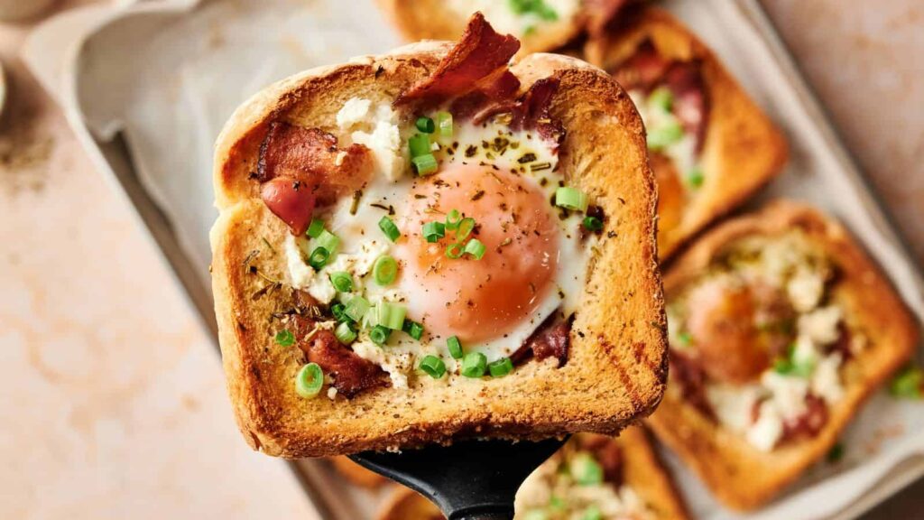A slice of toasted bread with a cooked egg in the center, topped with bacon, green onions, and herbs, held above a tray of similar pieces.