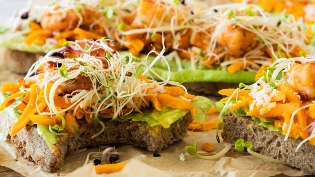 Close-up of open-faced sandwiches topped with shredded carrots, greens, sprouts, and chunks of cooked meat on a brown paper surface.