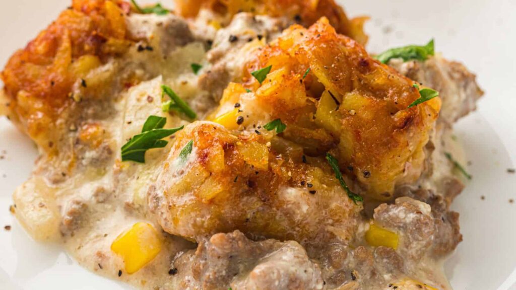 A close-up of a casserole dish featuring tater tots, creamy sauce, ground beef, corn, and garnished with chopped parsley.