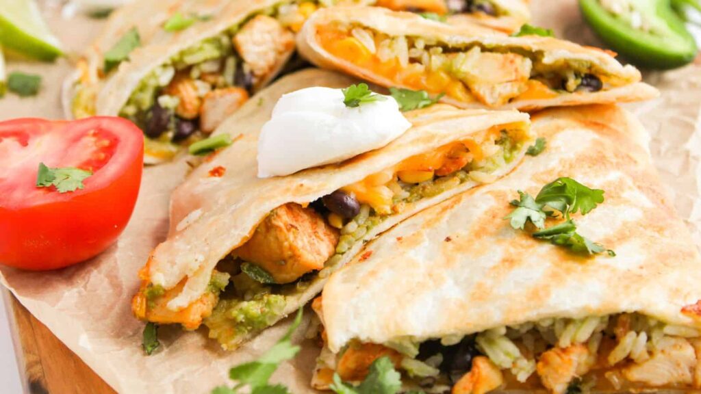 Close-up of a platter with quesadillas filled with chicken, rice, beans, cheese, and topped with sour cream. Garnished with cilantro, with sliced jalapeño and tomato on the side.