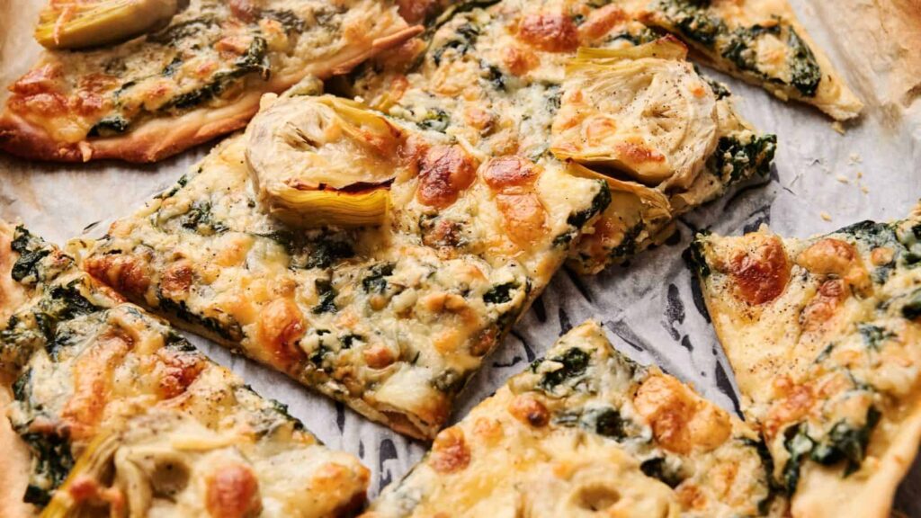Rectangular slices of flatbread pizza topped with melted cheese, spinach, and artichoke hearts on a parchment-lined surface.