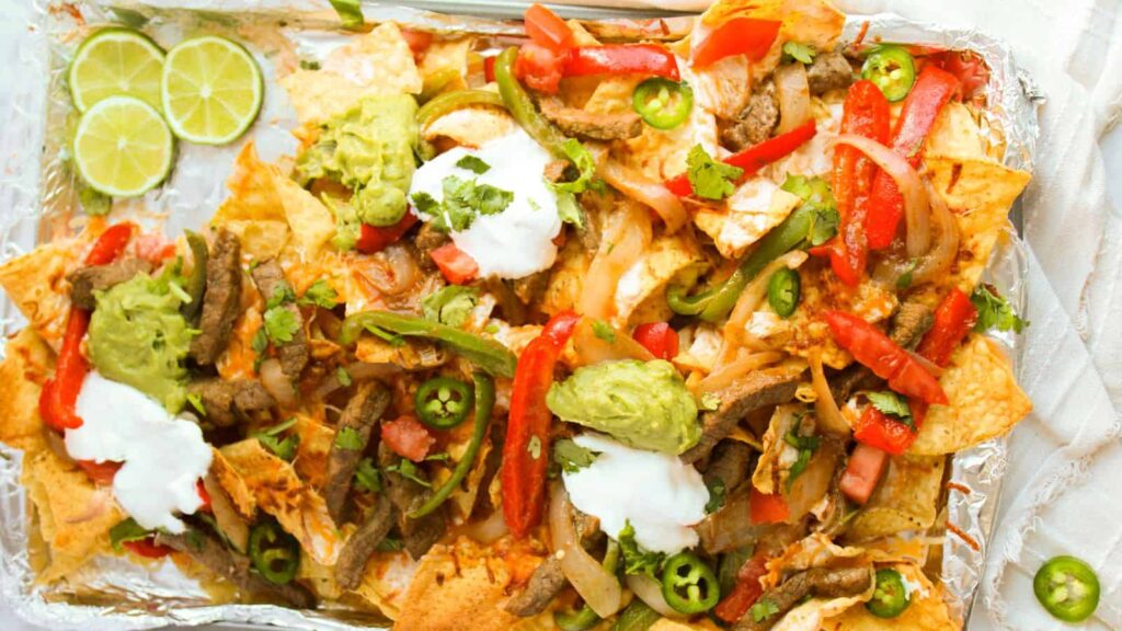 A tray of nachos with sliced beef, bell peppers, jalapeños, guacamole, sour cream, and lime slices.