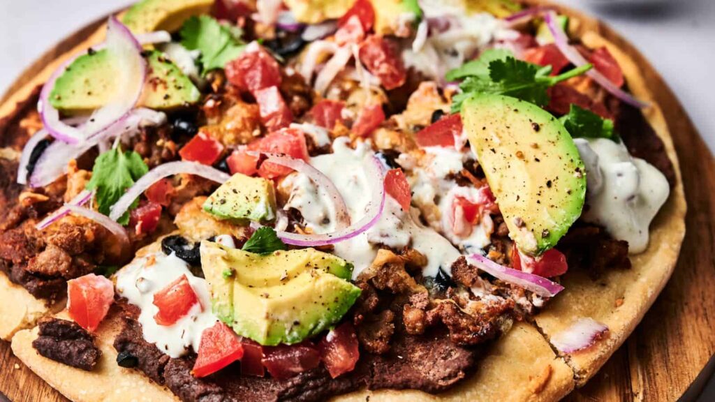 A loaded pizza topped with avocado slices, ground meat, black beans, diced tomatoes, red onion, fresh cilantro, and a drizzle of creamy white sauce.