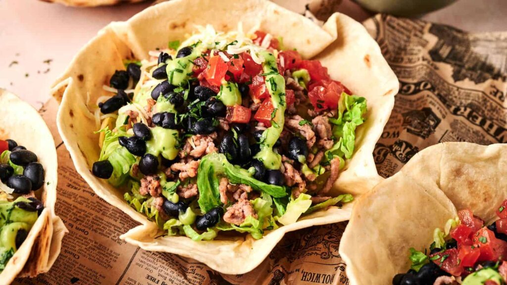 A taco salad served in a crispy tortilla bowl, topped with ground meat, lettuce, black beans, diced tomatoes, shredded cheese, and avocado dressing.
