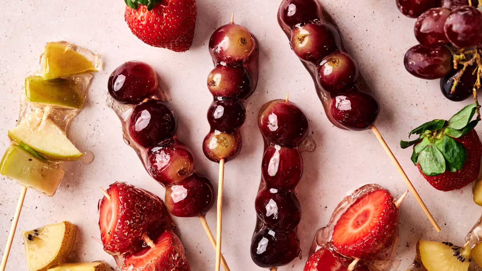 Skewers of grapes and strawberries coated in a shiny glaze are arranged on a light surface with loose fruit pieces nearby.
