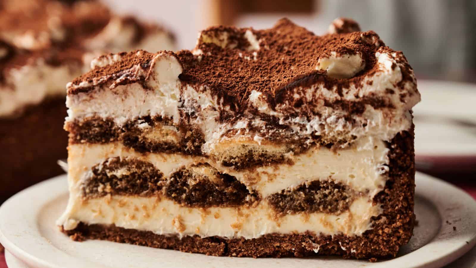 A close-up of a slice of layered tiramisu cake with cream, coffee-soaked sponge, and dusted cocoa powder on top, served on a white plate.