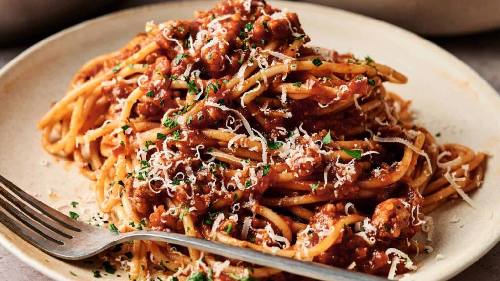 A plate of spaghetti topped with meat sauce, grated cheese, and chopped herbs, with a fork resting on the plate.