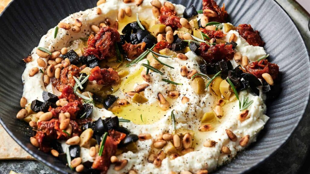 A bowl of whipped ricotta topped with olive oil, pine nuts, chopped black olives, sun-dried tomatoes, rosemary, and black pepper.