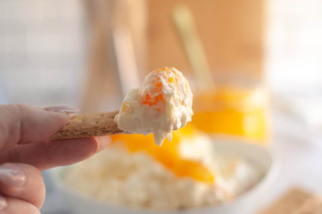 A hand holds a graham cracker topped with creamy Orange Fluff Salad Dip over a blurred bowl of the dip.