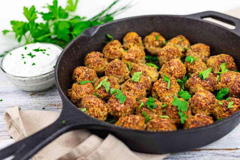A cast iron skillet filled with cooked meatballs garnished with fresh herbs, with a bowl of creamy dipping sauce and fresh parsley in the background.