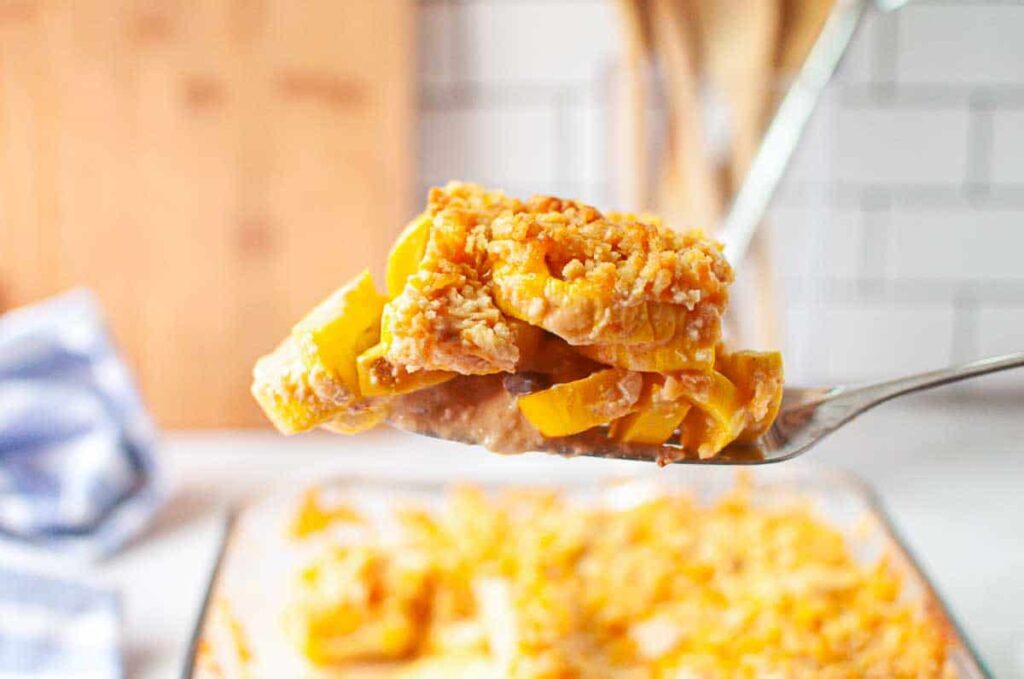 A metal spatula holds a serving of yellow squash casserole topped with a golden breadcrumb crust above a glass baking dish.