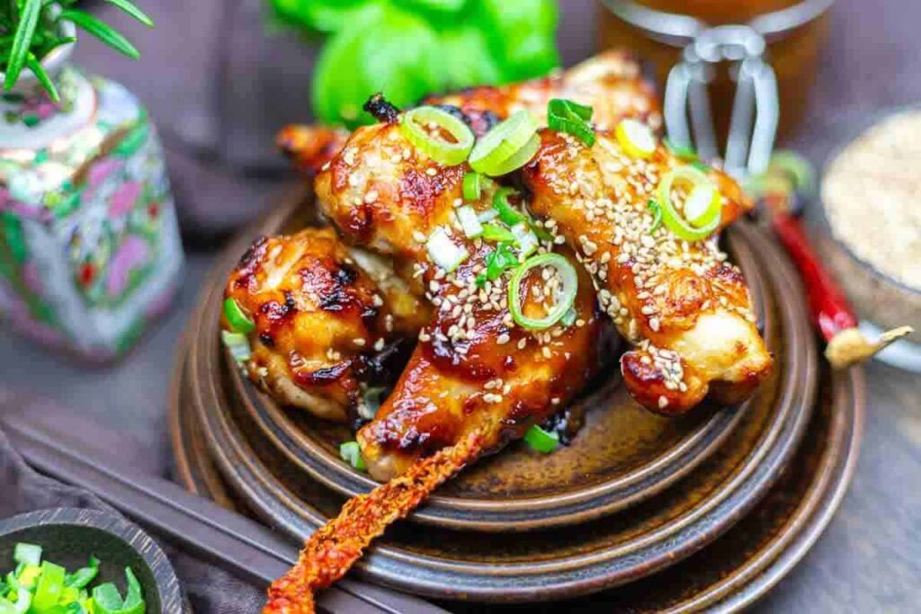 A plate of glazed chicken wings topped with sesame seeds and chopped green onions, garnished with a dried chili pepper.
