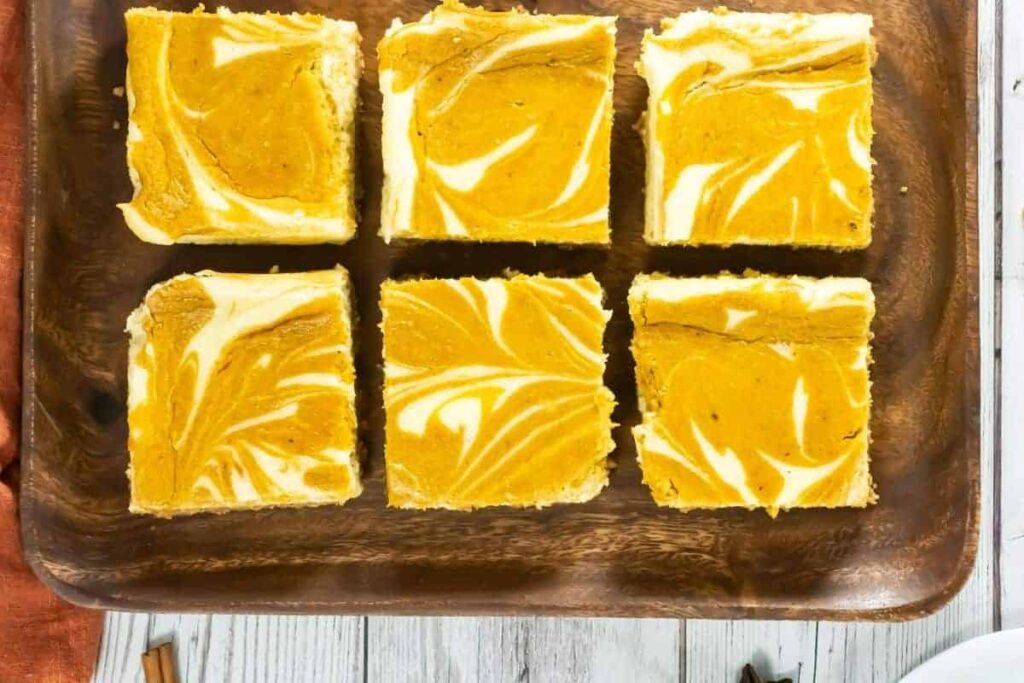 Six square pumpkin cheesecake bars are arranged on a wooden tray, displaying marbled orange and white patterns on top.