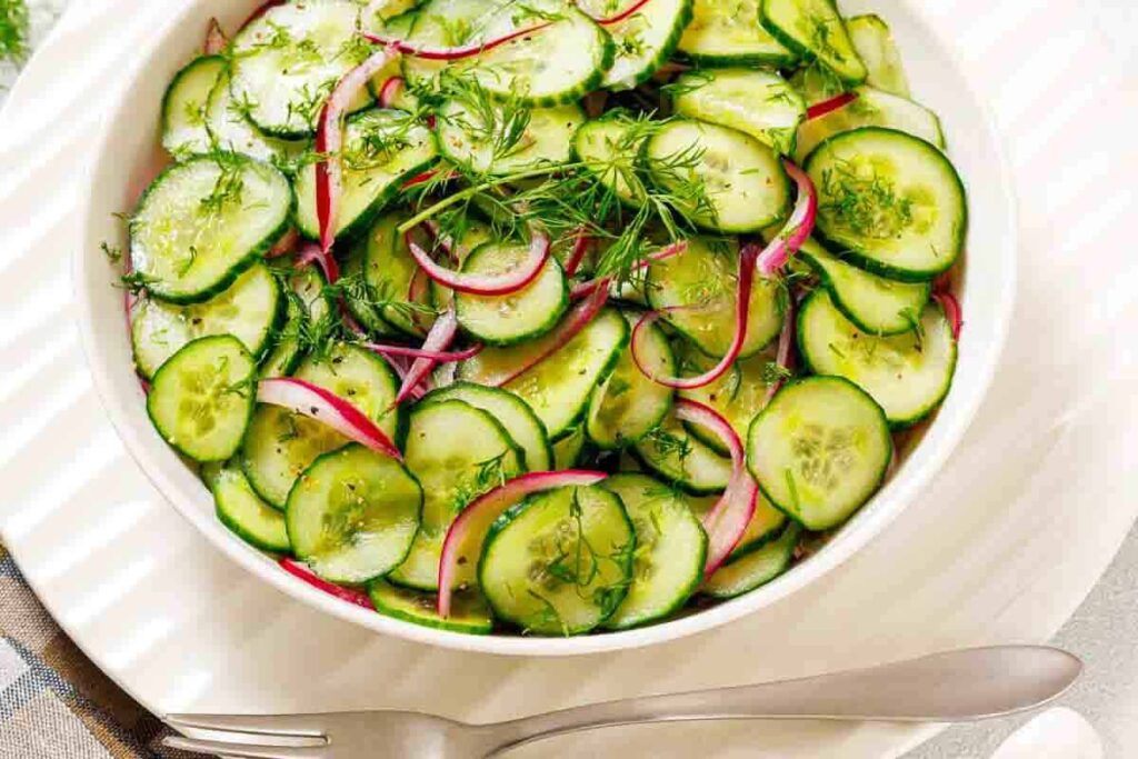 A bowl of cucumber salad with red onion slices and fresh dill, placed on a white plate with a fork and spoon beside it.